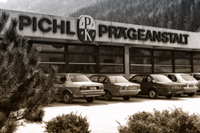 Historische Aufnahme: Firmengebäude 'Pichl Prägeanstalt' mit geparkten Autos, Glasfassade und Bergen im Hintergrund.